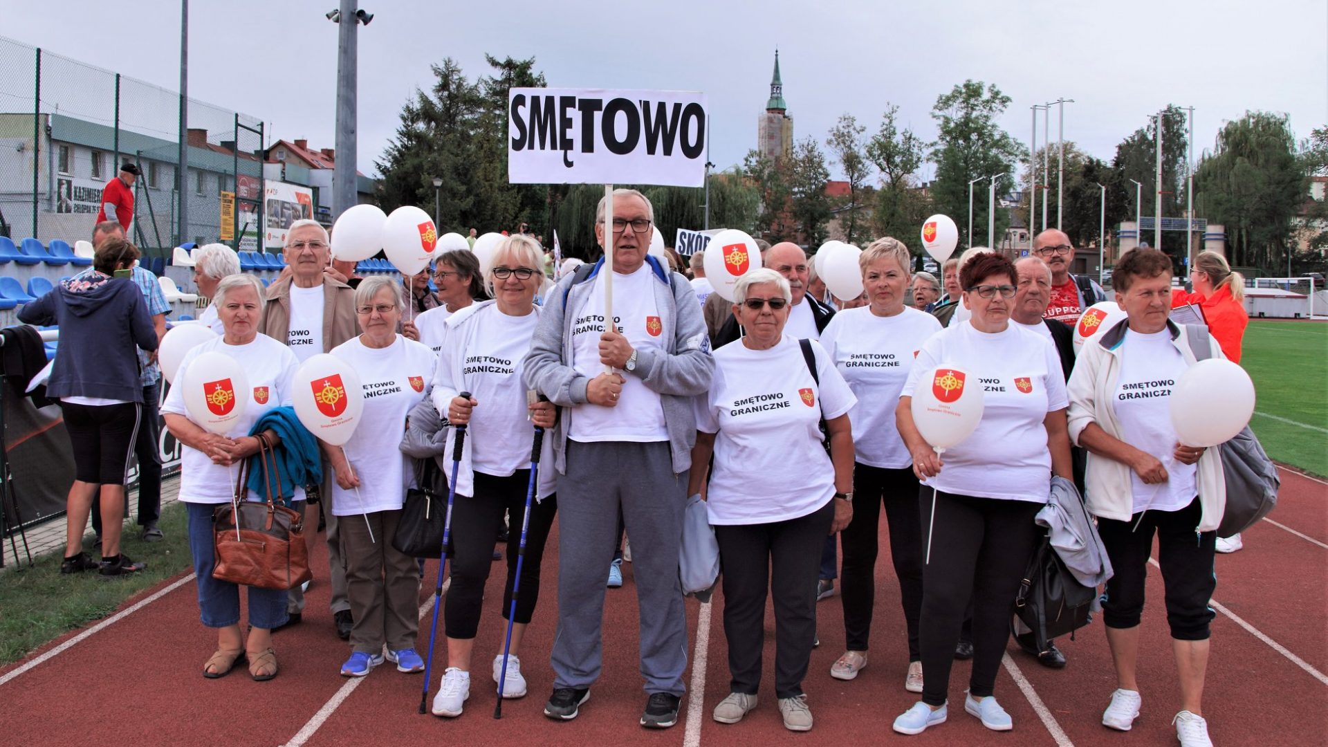 Członkowie klubu seniora ubrani na sportowo w koszulki z logo gminy Smętowo Graniczne stojący na bieżni stadionu